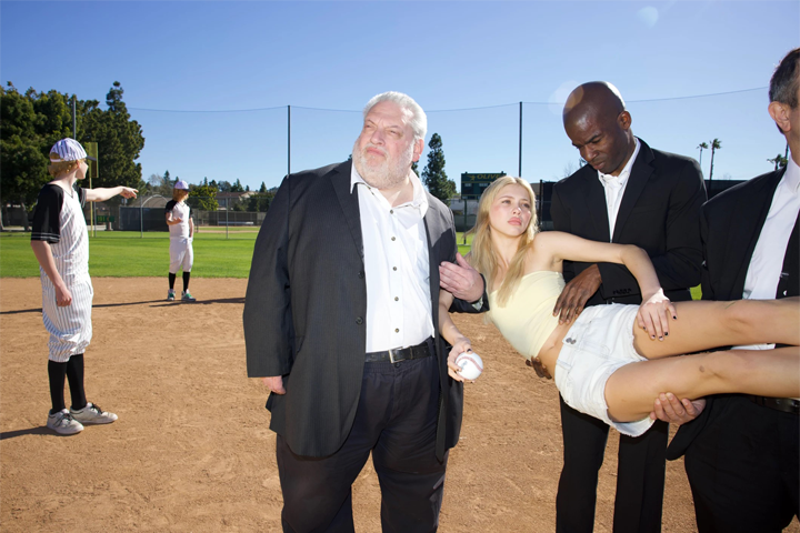 A young woman is held aloft by three men in suits while standing on a baseball field